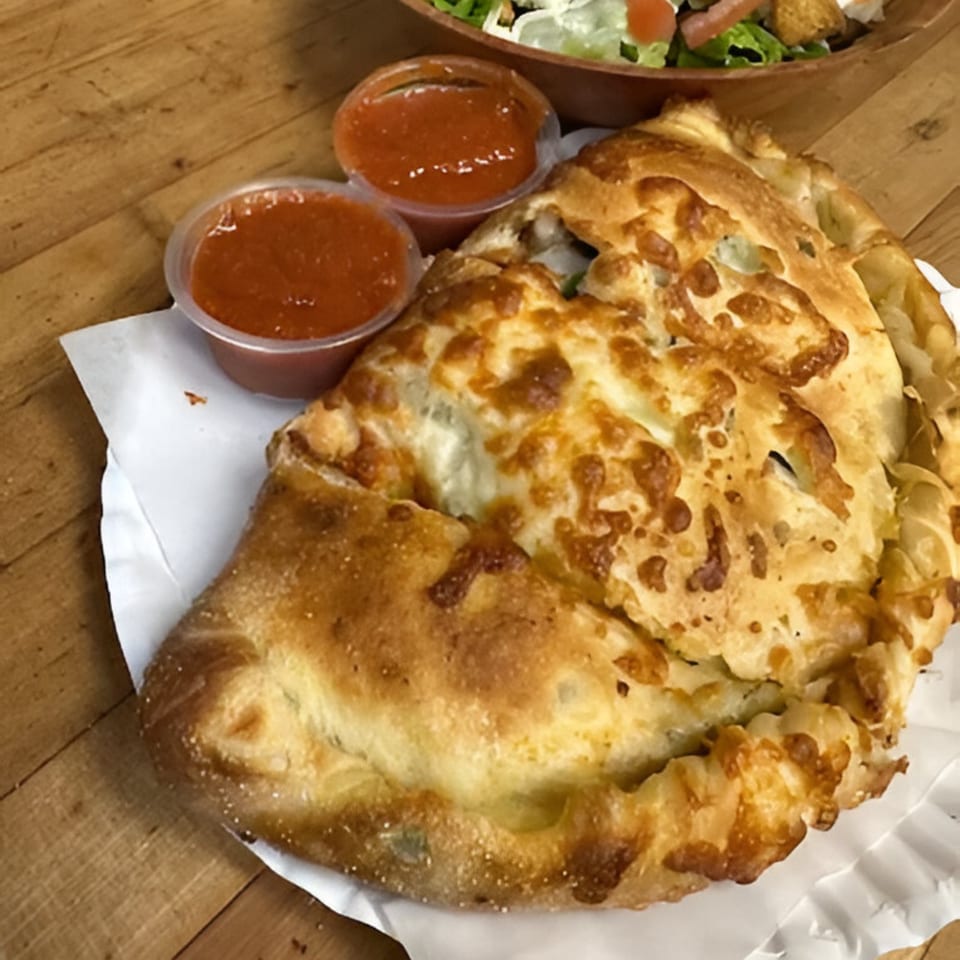 A golden-brown calzone with two cups of marinara sauce and a salad on a wooden table.